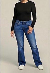 Femme portant un haut noir à manches longues, un jean bootcut bleu et des baskets blanches, debout avec une main dans la poche sur fond beige.