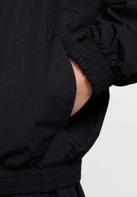 Svart vindjakke med elastiske mansjetter og midjebånd, med en sidelomme og en glatt, lett stofftekstur.