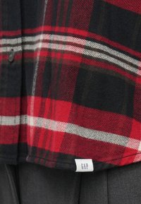 Czerwona i czarna flanelowa koszula w kratę z białymi i szarymi paskami. Posiada zapinany na guziki fason oraz małą metkę GAP na dole.