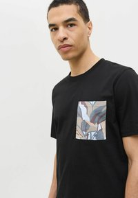 Sort t-shirt med korte ærmer lavet af bomuld, der har en rektangulær lomme med et flerfarvet bladmønster. Generelt glat tekstur.