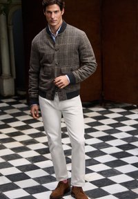 Chaqueta bomber de cuadros marrones con cuello de canalé negro, pantalones blancos y zapatos de ante marrones. Camisa azul visible debajo de la chaqueta.