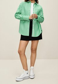 Osoba ubrana w zieloną i białą w paski koszulę zapinaną na guziki, czarną dżinsową spódnicę, białe skarpetki z grafiką oraz białe sneakersy sięgające za kostkę, stojąca na tle jednolitego tła.