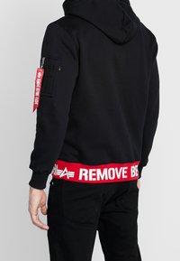 Felpa nera con cappuccio e tasca con zip sulla manica sinistra, presenta un patch rosso "RIMUOVI PRIMA DEL VOLO" sulla vita, tessuto testurizzato.