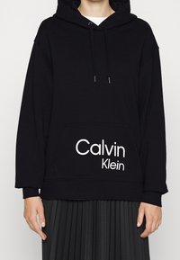 Osoba v černé mikině Calvin Klein s přední kapsou a bílým logem, v kombinaci s černou skládací sukní.