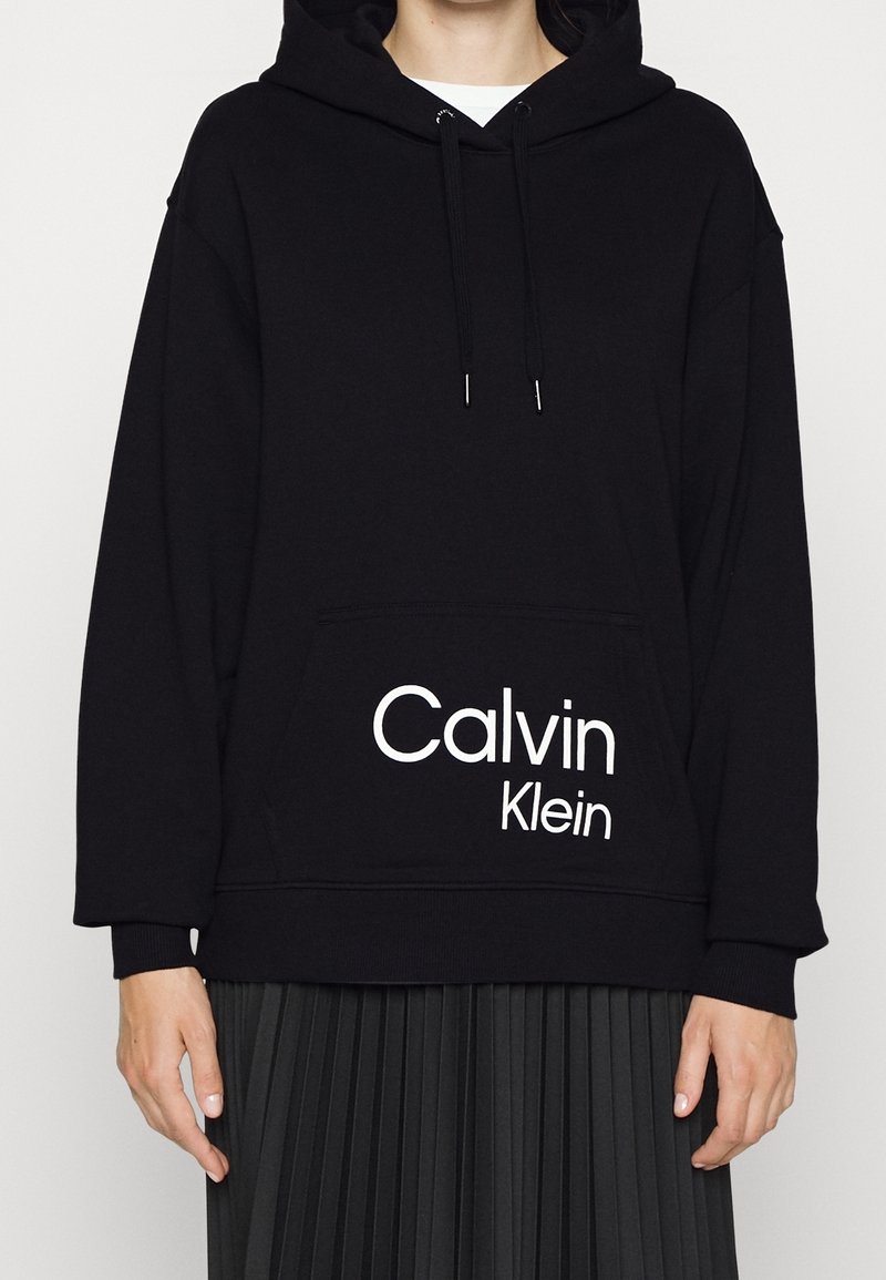 Osoba v černé mikině Calvin Klein s přední kapsou a bílým logem, v kombinaci s černou skládací sukní.