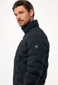 Černá péřová bunda s vysokým límcem, zipem vpředu a texturovanou látkou. Obsahuje malé logo na levém rukávu.