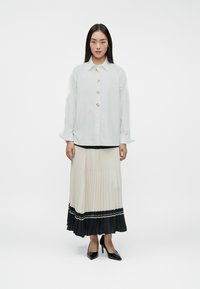 Camisa blanca con botones dorados, falda plisada en beige y negro, y tacones altos negros. Tela suave y diseño ajustado.
