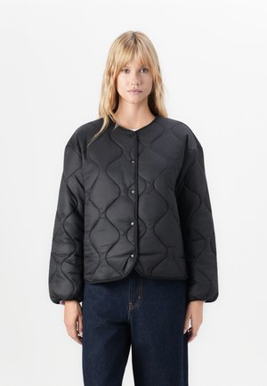 Blonde vrouw in een zwart gewatteerd button-up jasje en donkerblauwe jeans, staande tegen een witte achtergrond.