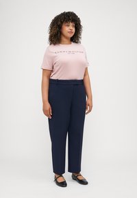 Helles rosa T-Shirt mit "TOMMY HILFIGER EST. 1985" in dunkelblau bedruckt. Marinefarbene Anzughose mit geradem Bein. Schwarze flache Schuhe.