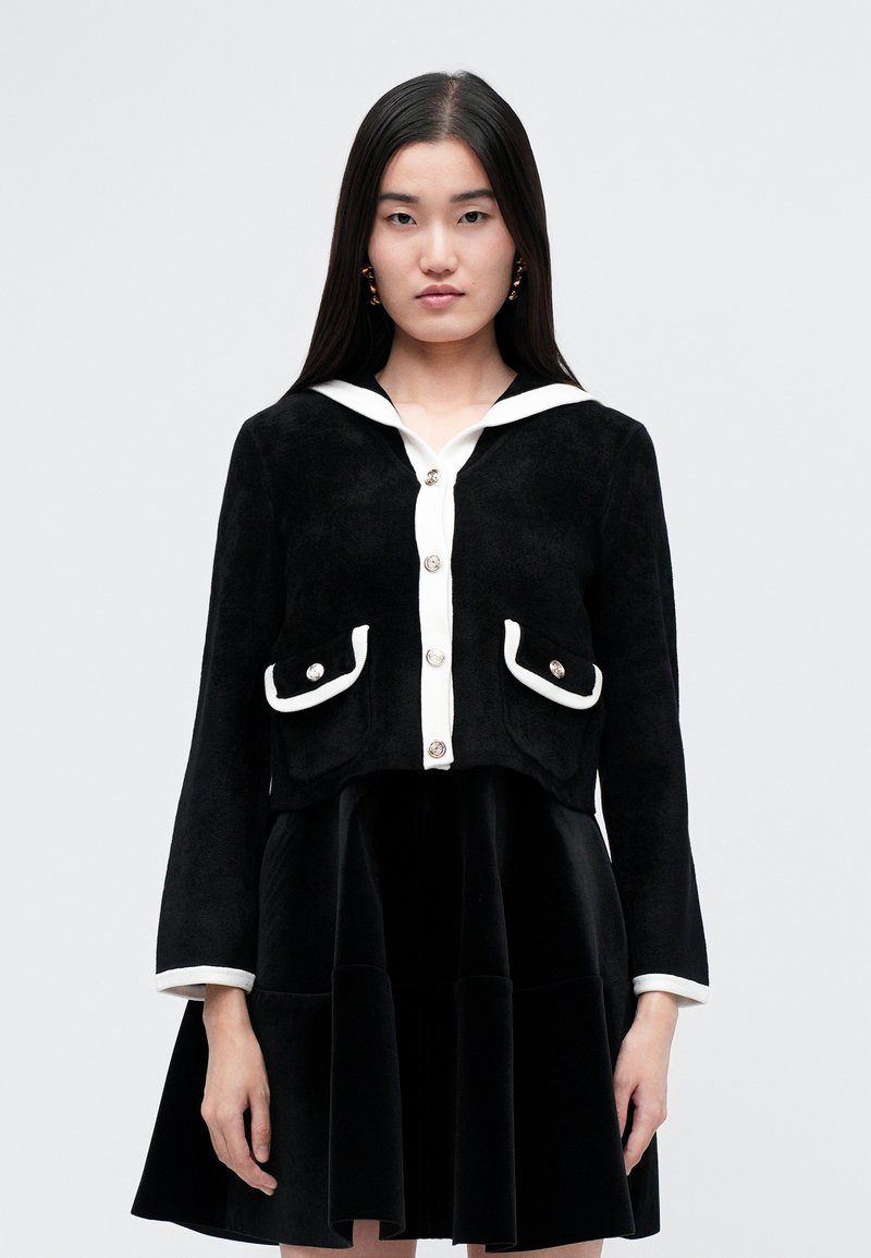 Schwarze, cropped Jacke mit weißem Kragen, Knopfverschluss und Fronttaschen. Texturiertes Material mit kontrastierenden Besatzdetails.