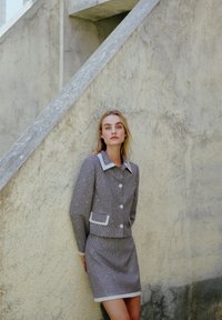 Completo houndstooth grigio e bianco con colletto, caratterizzato da bottoni e tasche. Texture liscia, design aderente e rifiniture a contrasto.