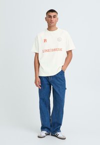 Off-white bomulds t-shirt med orange grafisk print og logo, parret med lige ben blå denimjeans og grå sneakers med sorte striber.