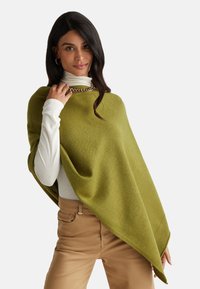 Poncho verde lavorato a maglia con una texture liscia, appoggiato su un collo alto bianco. Collana a catena notevole e pantaloni marrone chiaro visibili.