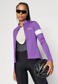 Jaqueta de ciclismo roxa com uma faixa branca, tecido leve, fecho completo e um logotipo. Calções de ciclismo pretos e um capacete preto segurados na mão.