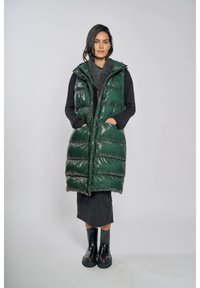 Długa, puszysta zielona kamizelka z połyskującym wykończeniem, z kapturem i bocznymi kieszeniami, narzucona na ciemny strój i zestawiona z czarnymi gumowymi butami.