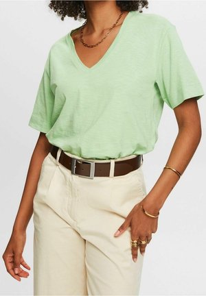 Person iført en lysegrøn V-hals t-shirt, stoppet ned i cremefarvede bukser med et brunt bælte, accessoriseret med guldringe, armbånd og en kædehalskæde.