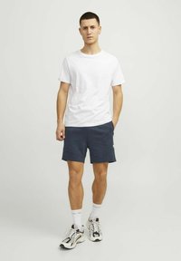 Witte T-shirt met ronde hals, gecombineerd met donkerblauwe shorts met zakken. gedragen met wit en zwart sport schoenen en witte sokken.
