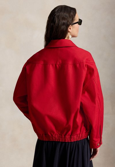 Femme aux longs cheveux foncés portant des lunettes de soleil noires et une veste rouge à taille élastique, vue de dos sur un fond beige uni.