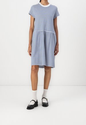 Femme portant une robe courte rayée bleu et blanc jusqu'aux genoux, des chaussettes blanches et des chaussures Mary Jane noires, debout contre un mur blanc.