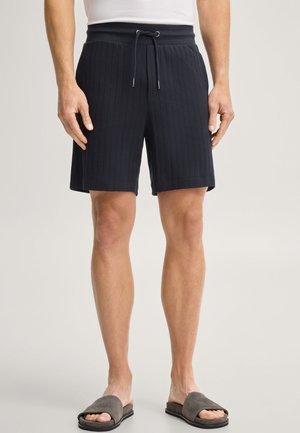Man draagt zwarte gestructureerde shorts met een trekkoord in de taille en grijze open teenslippers, staand tegen een effen achtergrond.