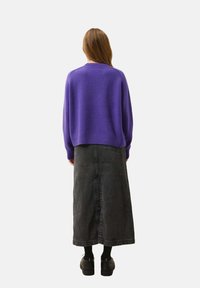 Lila gestrickter Pullover mit ribbeltem Halsausschnitt, kombiniert mit einem langen schwarzen Jeansrock. Schwarze Schuhe werden getragen, mit sichtbarer Stoffstruktur.