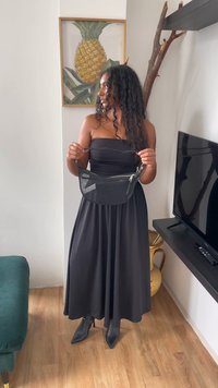 Zwarte mesh schoudertas met een gebogen vorm en een zilveren rits, vastgehouden door een persoon die een strapless zwarte jurk en spitse zwarte laarzen draagt.
