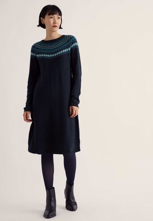 Femme portant une robe pull bleu marine foncé avec un empiècement à motifs bleus, des collants bleu marine et des bottines noires, debout devant un fond uni.