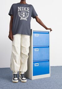 T-shirt graphique gris Nike, pantalon ample écru, et baskets montantes avec des accents noirs et jaunes, à côté d'un cabinet de classement bleu.