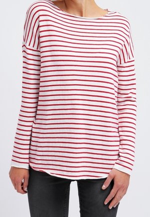 Femme portant une chemise blanche à manches longues avec des rayures horizontales rouges et un pantalon noir, debout, les bras détendus le long du corps.