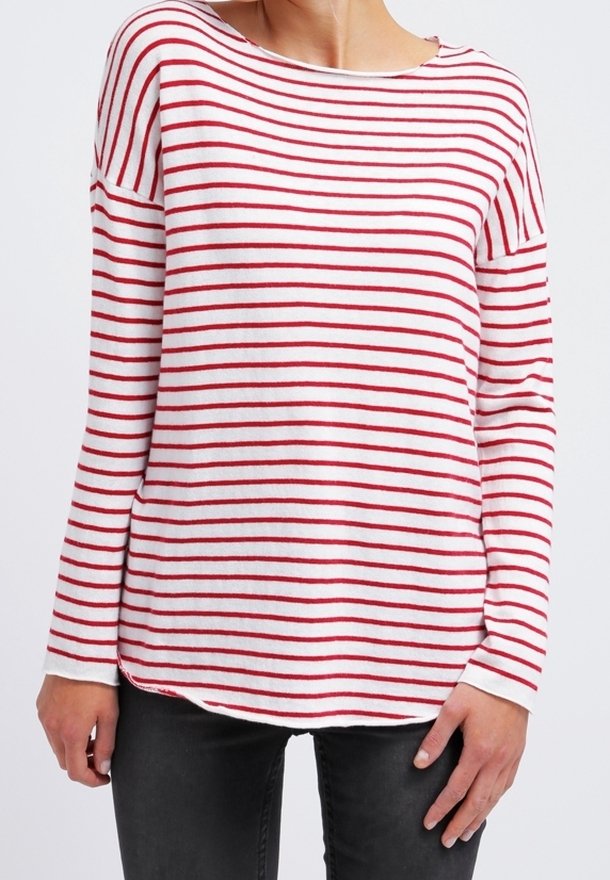 Femme portant une chemise blanche à manches longues avec des rayures horizontales rouges et un pantalon noir, debout, les bras détendus le long du corps.