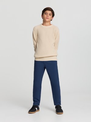 Ragazzo in piedi con le mani dietro la schiena, indossa un maglione lavorato a maglia color crema, pantaloni blu navy e sneakers nere su uno sfondo grigio chiaro uniforme.