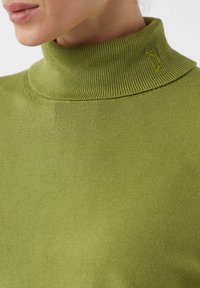 Zbliżenie na osobę w oliwkowym swetrze z golfem, z małym haftowanym logo w pobliżu kołnierza.