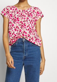 Kortärmad blus i ljusrosa med blommönster i nyanser av magenta. Mjuk tyg med rynkor vid halsringningen; bärs tillsammans med blå jeans.