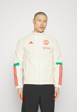 White zip-up jacket with red Adidas stripes on sleeves, Manchester United logo, and teal accent. Lightweight, smooth texture. Black pants below.