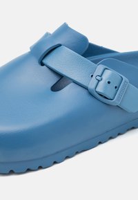 Zuecos de goma azul claro con una correa ajustable, superficie texturizada, punta abierta y suela estriada para mayor agarre. Incluye detalle de hebilla.