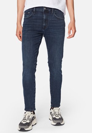 Homme portant un jean slim bleu foncé et des baskets épaisses noires et blanches, debout devant un fond blanc.