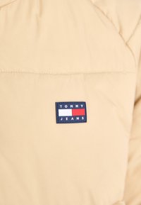 Beżowa kurtka puchowa z prostokątną granatową metką z napisem "TOMMY JEANS" oraz kolorowymi blokami czerwonym, białym i niebieskim. Miękka, gładka tekstura.