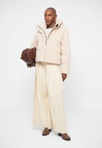 Puffy cropped cream jacket with hood, large front pockets, paired with wide cream trousers and brown heeled ankle boots. Holding a brown bag.