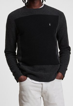 Mand iført mørkegrå sweater med sort firkantet frontpanel og lille hvid gedelogo, parret med lyse bukser. Hænder i lommerne.