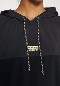 Černá mikina s kapucí, která má strukturovanou horní část, dva světle zbarvené značkové loga a kontrastní šňůrky s vzorovaným designem.