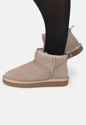 Stivali alla caviglia in suede beige con una soffice fodera in pelliccia, suola in gomma piatta e cuciture decorative. Presentano un risvolto arrotolato e una linguetta sul retro.