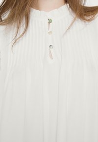 Blouse blanche avec des plis verticaux et trois boutons irisés près du col, portée par une personne aux longs cheveux bruns.