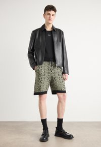 Chaqueta de cuero negra sobre una camiseta negra, combinada con pantalones cortos verdes con estampado, calcetines negros y zapatos negros. Los pantalones cortos tienen una cintura con cordón.