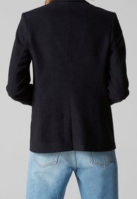 Blazer bleu marine foncé avec une coupe ajustée, tissu lisse et structure d'épaule subtile. Associe à un jean en denim bleu clair, avec poches arrière.