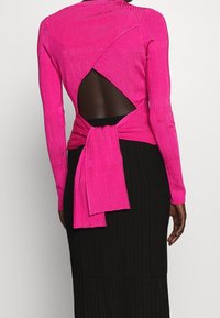 Langärmeliges Oberteil in leuchtendem Pink mit gerippter Struktur und einem offenen Rücken mit einer gebundenen Verzierung. Getragen über einem schwarzen, gerippten Rock.