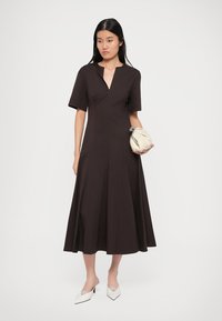 Καφέ, φόρεμα μέχρι το γόνατο με κοντά μανίκια και V-λαιμόκοψη. Φαρδύς φούστα και απαλό υλικό. Συνδυάζεται με μια ελαφριά τσάντα και λευκά τακούνια.