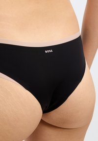 Melnas bikini stila apakšveļas biksītes ar gludu tekstūru. Tām ir mīksta, kontrastējoša gaiša jostas daļa un balti drukāts logo.