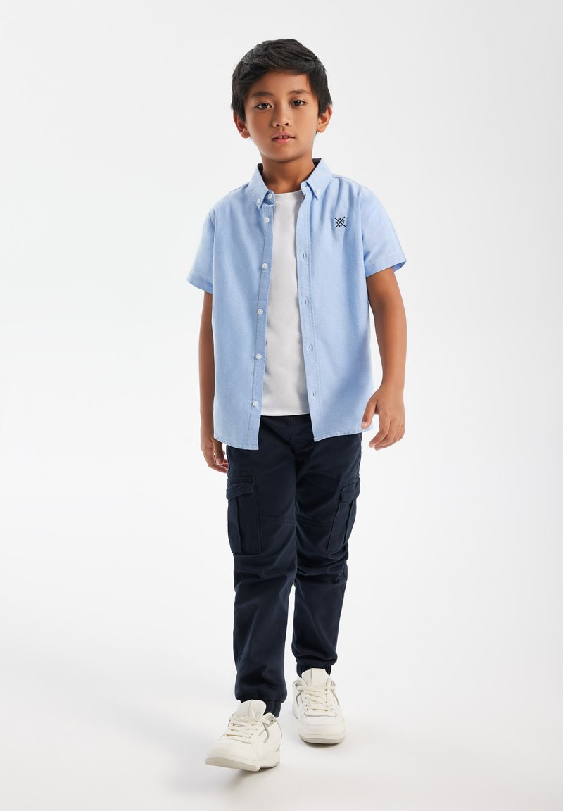 Poiss seisab, kandes helesinist lühikeste varrukatega nööpidega särki valge t-särgi peal, tumedaid lahtisi pükse ja valgeid tenniseid, valge tausta ees.