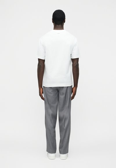 Homme debout, dos tourné, portant un t-shirt blanc uni à manches courtes, un pantalon gris et des baskets blanches, sur un fond blanc.