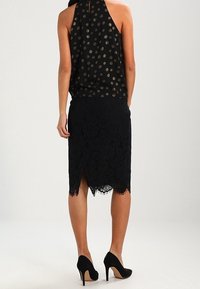 Femme vue de dos portant un haut sans manches noir à pois dorés, une jupe crayon en dentelle noire avec une fente à l'arrière, et des talons hauts noirs.